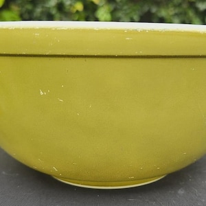 Puede incluir: Un cuenco mezclador verde lima vintage con un borde blanco y una fina línea de acento marrón oscuro. El cuenco tiene forma redondeada y se asienta sobre una base blanca. El cuenco es probablemente de cerámica o vidrio.
