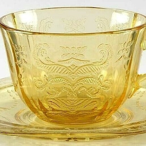 May include: A yellow glass teacup and saucer set with an intricate floral design. The set is made of clear glass with a yellow tint.