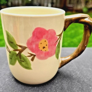 May include: Cream-colored ceramic mug with a pink floral design and a brown, branch-like handle. The flower has a yellow center, and green leaves and stems are also painted on the mug. A decorative piece for the kitchen.