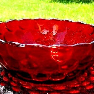 Puede incluir: Un cuenco de vidrio rojo brillante con un borde festoneado. El cuenco tiene una superficie texturizada y proyecta una sombra estampada. El cuenco es probablemente para servir o exhibir.