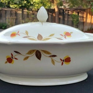 Puede incluir: Fuente de servir crema con tapa y diseño floral. La fuente tiene un borde festoneado, dos asas y una base de pedestal. La tapa está rematada con un adorno blanco en forma de lágrima. El patrón floral incluye hojas y flores naranjas, amarillas y marrones.