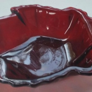 May include: A ruby red glass dish with a unique, organic shape. The dish has a wavy, scalloped edge and a glossy finish. The color is a deep, rich red, and the dish appears to be made of a thick glass material. This decorative dish could be used for serving or display.