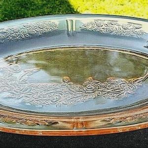 May include: Oval pink-tinted glass serving platter with embossed floral patterns. The platter has a slightly raised rim and two built-in cigarette rests. The floral design is repeated around the rim and in the centre.