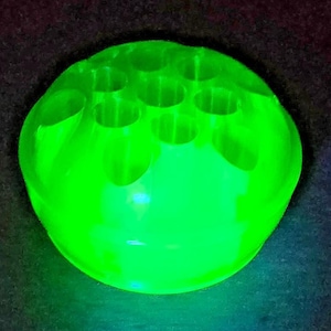 May include: A vibrant, luminous green glass flower frog with multiple holes. The round, bowl-shaped frog is made of a translucent material and sits on a dark surface. The holes are evenly spaced across the top.