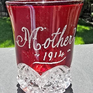 以下が含まれることがあります： カットグラスの底を持つ濃い赤色のガラスのタンブラー。グラスには「Mother - 1914」とエッチングされています。