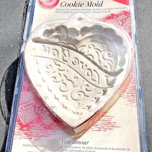 Puede incluir: Un molde para galletas de corazón blanco con las palabras "True Love" y "I Love You" en relieve. El molde está empaquetado en una bolsa de plástico transparente con un fondo rojo y blanco. El empaque también incluye las palabras "Ovenproof!" y "Vrai amour".