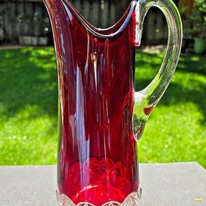 May include: A tall, ruby red glass jug with a clear glass handle. The jug has a decorative, cut-glass base. The handle is arched and attached to the top and side of the jug. The jug is on a dark surface with a green background.