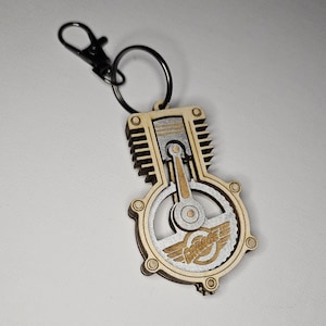 May include: A wooden keychain shaped like a cutaway engine with a silver finish. The engine has a silver gear and a silver piston. The keychain has a black metal ring and a silver logo that says "Porsche" with wings.