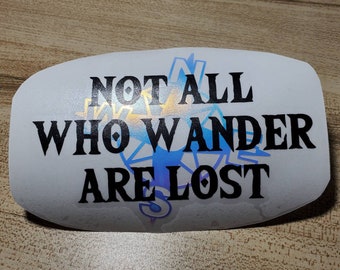 May include: White sticker with the text "NOT ALL WHO WANDER ARE LOST" in black, with a blue and gold compass design in the background. The sticker has rounded edges and is on a wooden surface.