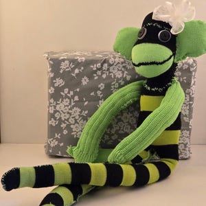 May include: A handmade sock monkey toy crafted from black and lime green striped fabric. The monkey features two dark button eyes, a light green face, and a white ribbon bow attached to the top of its head. It has long arms and legs.