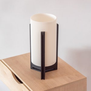 May include: A white cylindrical lampshade with black vertical slats sits on a wooden bedside table. The lampshade is on a black metal stand with a geometric design.