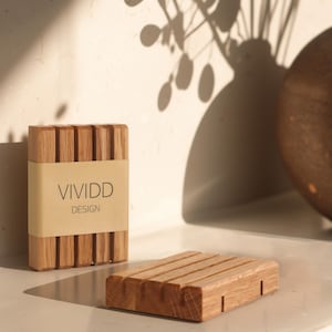 Puede incluir: Juego de jabonera y soporte para jabón de madera. La jabonera es rectangular con ranuras paralelas. El soporte para jabón es un bloque rectangular con ranuras verticales y una etiqueta que dice "VIVIDD DESIGN". El juego está hecho de madera clara.