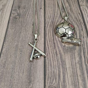 May include: Two silver-toned necklaces. One features a baseball and crossed bats pendant. The other has a soccer ball and cleat pendant. Both pendants hang from a delicate chain, perfect for sports enthusiasts.