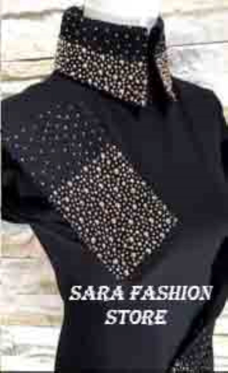May include: Black shirt with a rhinestone design on the collar and a diagonal panel on the front. The shirt has the text "SARA FASHION STORE" printed on the front.
