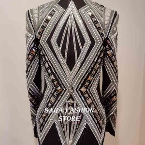 Op de afbeelding: Een zwart blazer met een geometrisch ontwerp met zilveren en witte versieringen. De blazer heeft een hoge kraag en lange mouwen. De tekst "SARA FASHION STORE" is zichtbaar op de voorkant van de blazer.