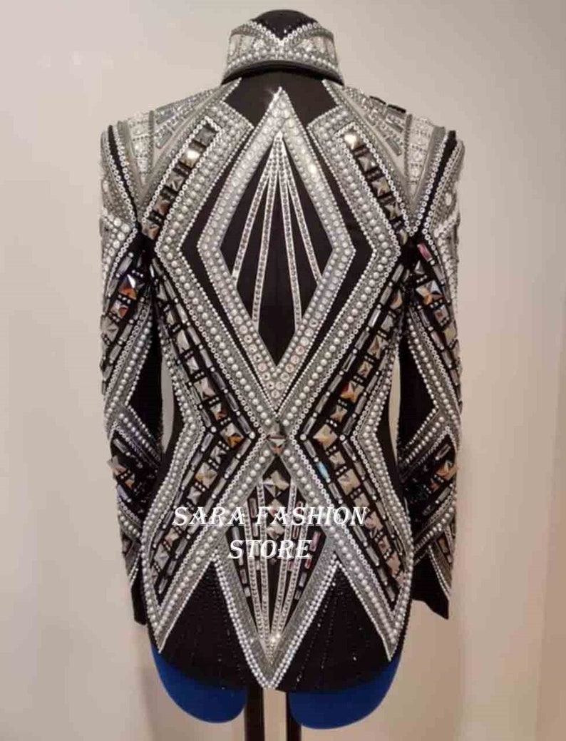 Op de afbeelding: Een zwart blazer met een geometrisch ontwerp met zilveren en witte versieringen. De blazer heeft een hoge kraag en lange mouwen. De tekst "SARA FASHION STORE" is zichtbaar op de voorkant van de blazer.