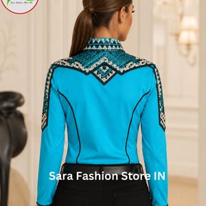 May include: A bright turquoise long-sleeve shirt featuring intricate black and silver geometric crystal embellishments across the shoulders, collar, and cuffs. The shirt includes black piping accents and is paired with black pants and a black belt.