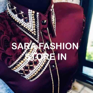 Könnte beinhalten: Ein burgunderfarbenes Satinhemd mit einem Spitzenkragen und -manschetten. Das Hemd ist mit goldenen und silbernen Strasssteinen verziert und der Text "SARA FASHION STORE IN" ist auf der Vorderseite aufgedruckt.