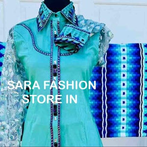 May include: A turquoise blue shirt with a lace overlay and rhinestone embellishments. The shirt has a western style collar and a button-up front. The text "SARA FASHION STORE IN" is visible on the shirt. The shirt is displayed with a blue and white patterned fabric.
