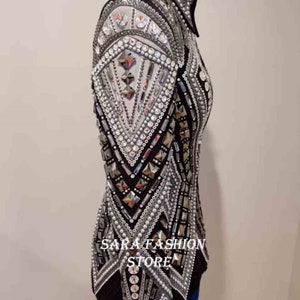 Op de afbeelding: Een zwart en zilveren showjasje met een geometrisch ontwerp en parel accenten. Het jasje heeft een hoge kraag en lange mouwen. De tekst "SARA FASHION STORE" is zichtbaar op de voorkant van het jasje.