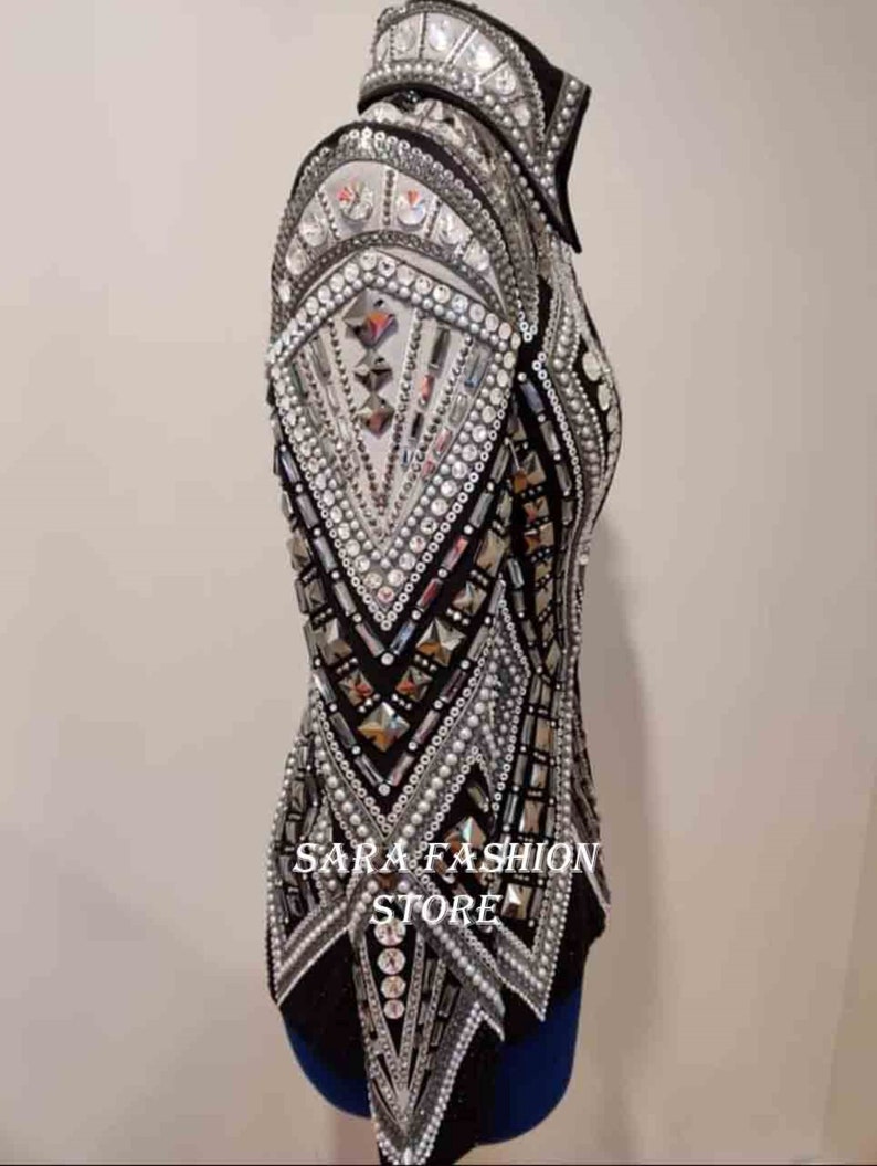 Op de afbeelding: Een zwart en zilveren showjasje met een geometrisch ontwerp en parel accenten. Het jasje heeft een hoge kraag en lange mouwen. De tekst "SARA FASHION STORE" is zichtbaar op de voorkant van het jasje.