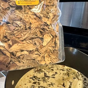 May include: A clear plastic bag filled with dried banana leaves. The bag has a yellow label with the text "Palankey Original Pinoy Goods" and website information. The leaves are being poured into a black pot with a creamy white sauce and green leafy vegetables.