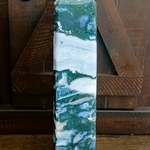May include: A green and white agate crystal point, also known as a green moss agate, is a natural stone with a unique swirling pattern. The crystal is polished and has a smooth, glossy finish.