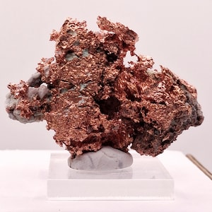 May include: A raw native copper mineral specimen featuring a metallic, reddish-brown, crystalline structure with jagged, textured surfaces and small patches of light grey matrix rock, mounted securely on a clear, square acrylic display base.