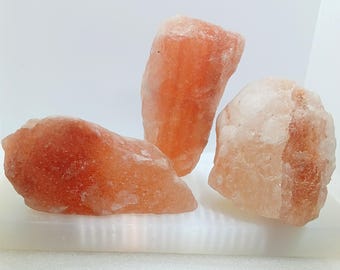 Puede incluir: Tres rocas de sal rosa del Himalaya en bruto. Los trozos de sal varían en tamaño y forma, con una gama de tonos rosados que van del claro al rosa intenso. Las rocas se exhiben sobre una superficie blanca.