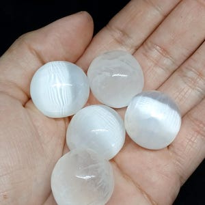 May include: Five white selenite spheres, each with a smooth, rounded shape. The spheres are held in an open hand, showcasing their translucent, almost pearlescent quality. The background is a dark, solid color.