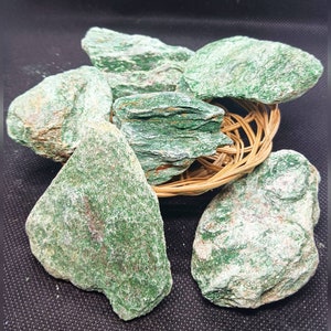 Puede incluir: Una colección de piedras verdes sin pulir con un aspecto moteado y en capas. Las piedras están dispuestas en una cesta tejida.