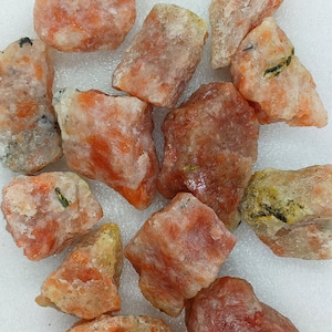 May include: A collection of rough, orange sunstone crystals with a translucent, glassy appearance. The crystals have a variety of shapes and sizes, and some have a slight green tint.