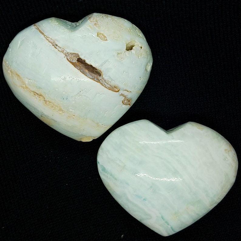 May include: Two heart-shaped stones in a pale blue-green colour with natural veining. One stone has a visible brown fissure. The hearts are displayed against a black background, highlighting their unique textures and colours. These are likely decorative items.