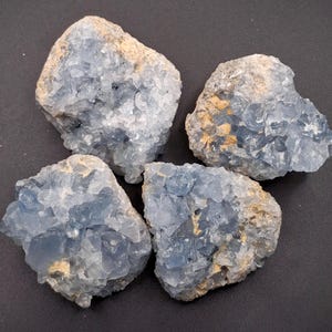 May include: Four raw celestite crystal clusters featuring pale blue, translucent, angular crystal formations embedded in a rough, tan-colored rocky matrix. Each piece displays a jagged, irregular shape with varying sizes of crystalline points.