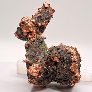 May include: A raw mineral specimen featuring a dark, textured, and craggy matrix with prominent, metallic, bright copper-coloured deposits. The structure has an irregular, organic shape with a vertical protrusion and a rounded base section.