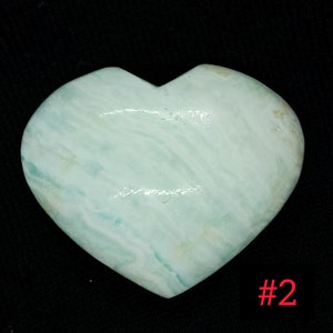 May include: A heart-shaped stone in shades of pale blue-green with white marbling. The polished stone is set against a black backdrop. The number "#2" is visible in the bottom right corner.