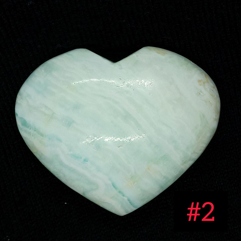 May include: A heart-shaped stone in shades of pale blue-green with white marbling. The polished stone is set against a black backdrop. The number "#2" is visible in the bottom right corner.