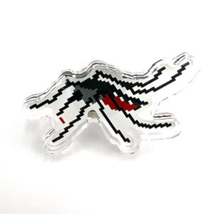 May include: A clear acrylic pin with a pixelated image of a white and black spaceship with red accents. The spaceship is flying in a diagonal direction.