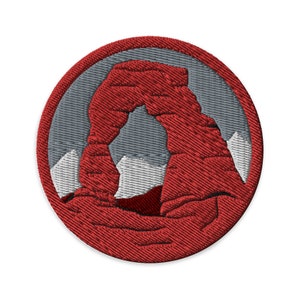 Pode incluir: Patch bordado com o Delicate Arch vermelho sobre um fundo cinza, circundado por uma borda vermelha. O design inclui formas de montanhas brancas. Adequado para jaquetas, bolsas ou outros artigos têxteis.