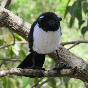 Könnte beinhalten: Ein handgefertigter, schwarz-weiß gestrickter Vogel, der auf einem Ast sitzt. Der Vogel hat eine weiße Brust, schwarze Flügel und einen Schwanz sowie kleine schwarze Beine. Der Vogel ist aus Garn gefertigt und wirkt realistisch.