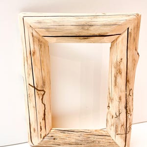 May include: A square wooden picture frame with a natural, light-coloured finish. The frame is constructed from thick, rough-hewn wood, showcasing the natural grain and texture. The corners are joined at right angles, creating a sturdy, rustic aesthetic.