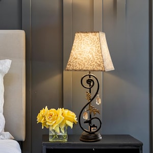 Op de afbeelding: Een decoratieve tafellamp met een patroon lampenkap en een zwarte, sierlijke voet. De voet heeft een wervelend ontwerp met gouden accenten en hangende kristalachtige ornamenten. Een vaas met gele rozen staat in de buurt.