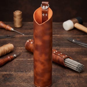 May include: A cylindrical brown leather case with a smooth, mottled texture, featuring a vertical strap secured by silver-toned metal rivets and a top ring for hanging. The case has a V-shaped opening at the top and visible stitching along the base.
