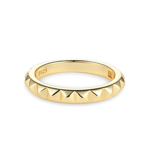 Op de afbeelding: Een goudkleurige ring met een noppenontwerp. De ring heeft een reeks kleine, piramidevormige noppen rond de band. De ring is gemaakt van glanzend, goudkleurig metaal en staat tegen een witte achtergrond.