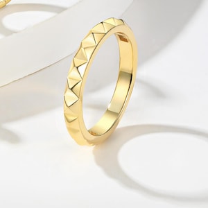 Peut inclure: Une bague dorée avec un motif géométrique de formes triangulaires sur le pourtour. La bague est posée sur un fond blanc, avec une ombre projetée sur la droite. Il s'agit d'un bijou.