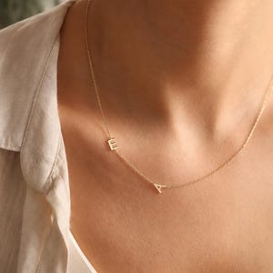 May include: A delicate gold-toned chain necklace featuring two small, uppercase letter charms, specifically the letters E and A, positioned asymmetrically along the thin metal links. The necklace rests against the neck, showcasing a minimalist design.