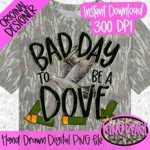 Puede incluir: Una camiseta con estampado de camuflaje gris que presenta un gráfico de una paloma volando y dos cartuchos de escopeta. El texto dice BAD DAY TO BE A DOVE. El diseño incluye letras negras con un contorno texturizado sobre un fondo de camuflaje.