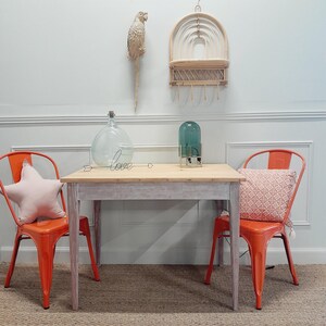 Op de afbeelding: Een witgeschilderde houten tafel met twee oranje metalen stoelen. De tafel heeft een draadwoord "love" erop. Er staat een glazen fles en een groene glazen lamp op de tafel. Een wit houten rek met een geweven ontwerp hangt aan de muur achter de tafel.