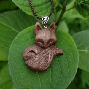 Op de afbeelding: Een houten vos-hangerketting met een bruin koord. De vos is gegraveerd in een opgerolde positie met zijn staart om zijn lichaam gewikkeld. De hanger hangt aan een groen blad.