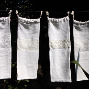 Puede incluir: Cuatro bolsas de lino blanco colgando de un tendedero. Cada bolsa tiene un delicado borde de encaje y está bordada con un diseño floral.
